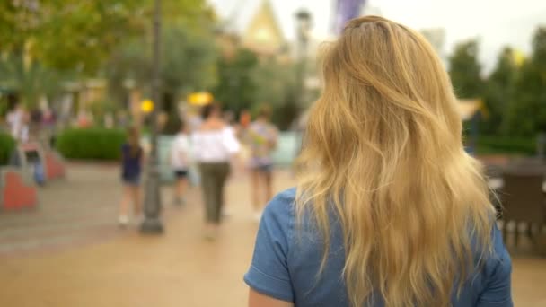 Eine schöne junge blonde Frau geht durch die Stadt und schaut sich interessiert um, die Kamera folgt ihr vor dem Hintergrund einer sich bewegenden Menschenmenge, verschwommen — Stockvideo