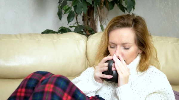 Donna malata in un maglione sotto una coperta che beve una bevanda calda sul divano in soggiorno — Foto Stock