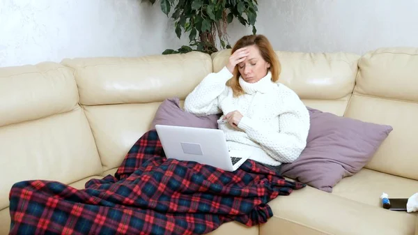 Mujer rubia con un frío sentado en el sofá en un suéter y una cuadros llamando a su médico en un ordenador portátil a través de enlace de vídeo . —  Fotos de Stock