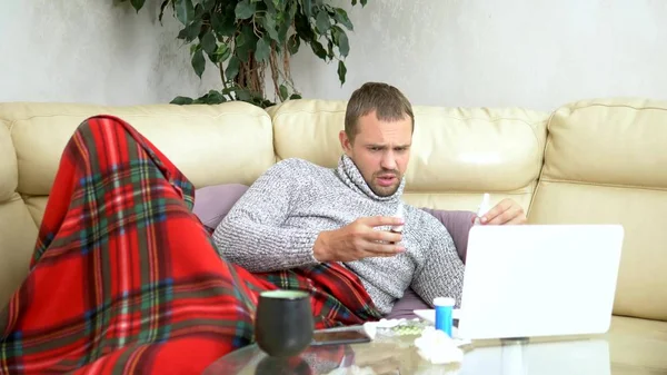 Homem com um frio sentado no sofá em uma camisola e uma xadrez chamando seu médico em um laptop via link de vídeo . — Fotografia de Stock