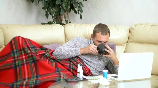 Homme avec un rhume assis sur le canapé dans un pull et un plaid appeler son médecin sur un ordinateur portable via lien vidéo . — Photo