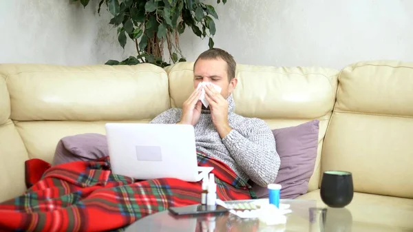 man with a cold sitting on the sofa in a sweater and a plaid calling his doctor on a laptop via video link.