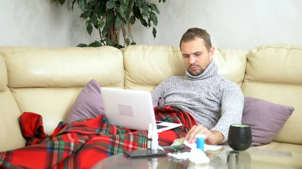 Homem com um frio sentado no sofá em uma camisola e uma xadrez chamando seu médico em um laptop via link de vídeo . — Fotografia de Stock