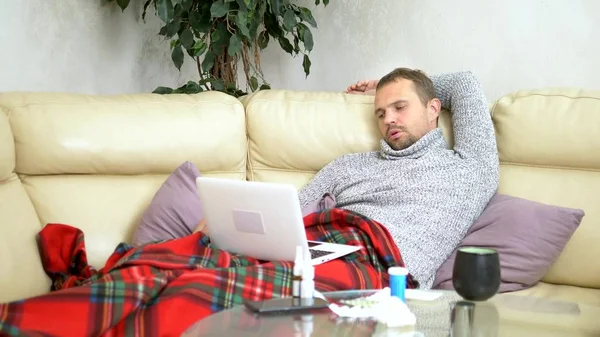 Hombre con un frío sentado en el sofá en un suéter y una cuadros llamando a su médico en un ordenador portátil a través de enlace de vídeo . —  Fotos de Stock
