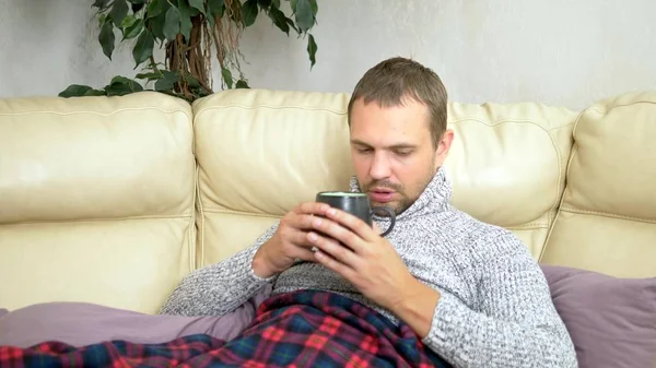 Mladý nemocný muž v svetru pod plénou s horečkou kontroluje teplotu s teploměrem doma — Stock fotografie