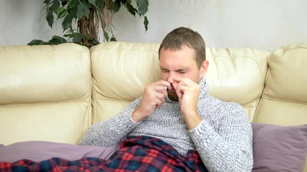 Tampan pria dengan hidung meler menggunakan semprotan hidung di rumah, duduk di sofa di ruang tamu di sweter dan di bawah selimut — Stok Foto