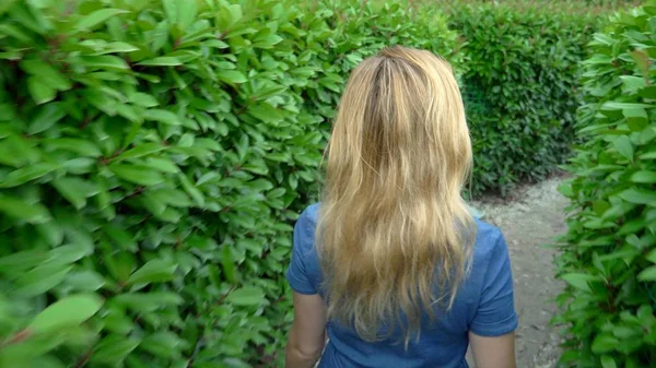 Seorang wanita dalam setelan denim pendek berjalan melalui labirin lindung pada hari musim panas yang cerah. tampilan belakang — Stok Foto