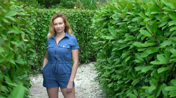 A woman in a short denim jumpsuit standing in the middle of a hedge maze on a sunny summer day. Looks at the camera. — Stock Photo, Image