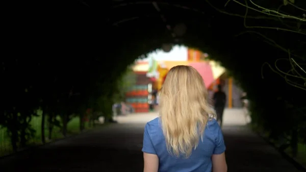 Eine blonde Frau mit langen Haaren geht durch einen dunklen Tunnel aus Kletterpflanzen. die Kamera folgt ihr. — Stockfoto