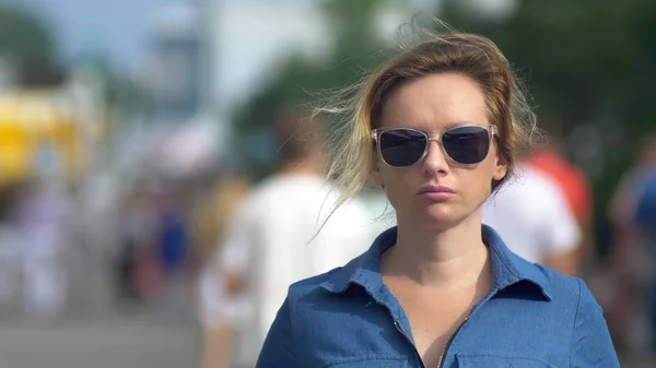 Beautiful woman in sunglasses, looking at camera, close-up. hot summer day. Moving crowd of people blurred in motion in the background. — Stock Photo, Image