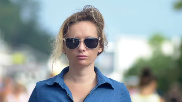 Hermosa mujer con gafas de sol, mirando a la cámara, de cerca. caluroso día de verano. Mucha gente en movimiento se desdibujó en el fondo . — Foto de Stock