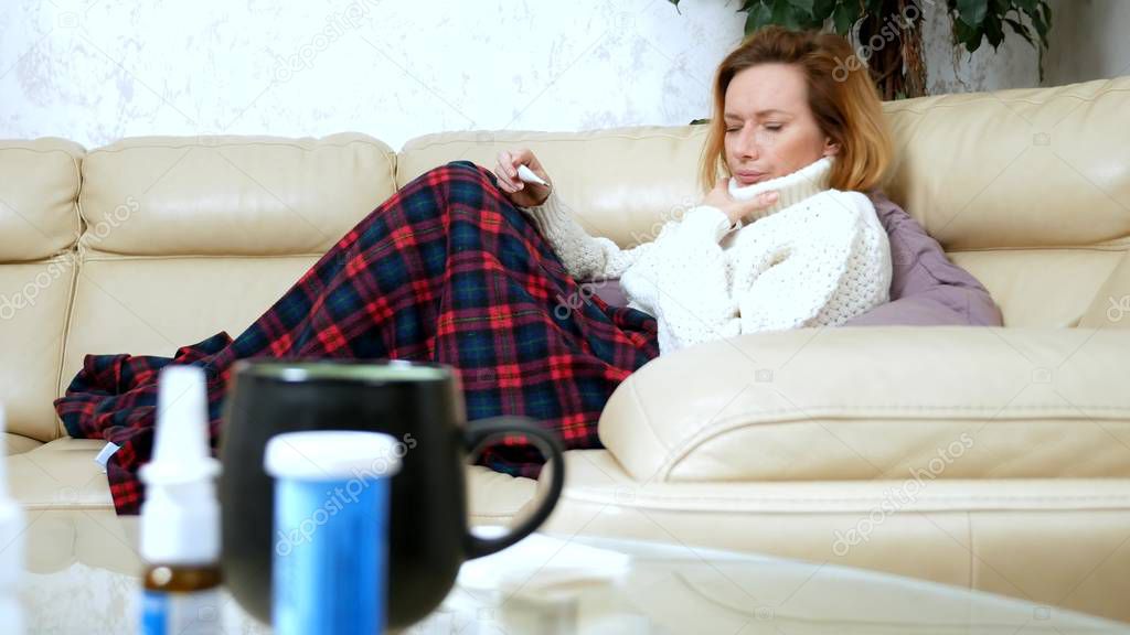 Young sick woman in a sweater under a plaid with a fever checks her temperature with a thermometer at home