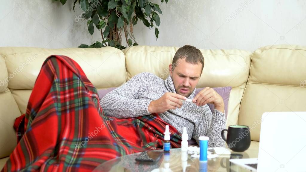 Young sick man in a sweater under a plaid with a fever checks his temperature with a thermometer at home