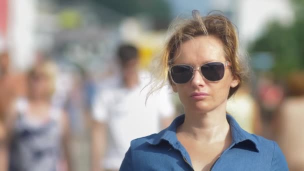 Schöne Frau mit Sonnenbrille, Blick in die Kamera, Nahaufnahme. Heißer Sommertag. Bewegende Menschenmenge im Hintergrund verschwommen. — Stockvideo