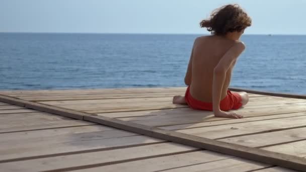 巻き毛を持つティーンエイジャーの少年は、海の上に木製のテラスに座っている海の風景を背景に海を見ます — ストック動画