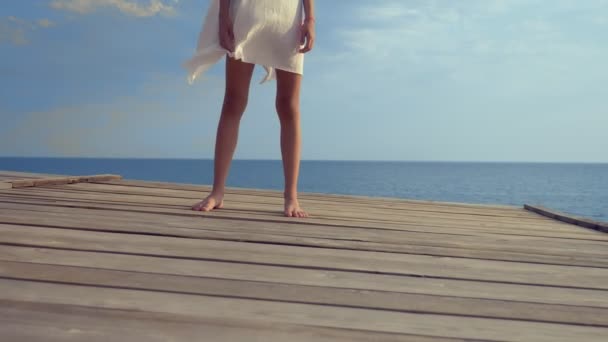 Menina adolescente em um vestido branco com cabelos longos olha pensativamente para o mar, estando em um terraço de madeira acima do mar. vento desenvolve cabelo — Vídeo de Stock