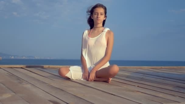 Teenager-Mädchen in weißem Kleid mit langen Haaren blickt nachdenklich ins Meer, auf einer Holzterrasse über dem Meer. Wind entwickelt Haare — Stockvideo