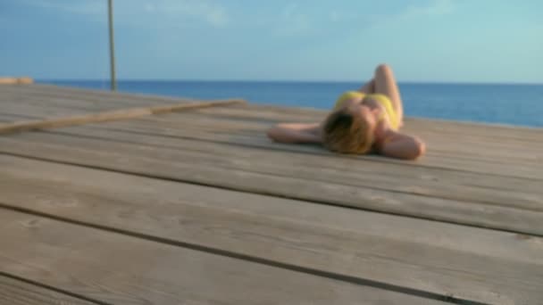 Schöne schlanke Frau mit wunderschönen Brüsten in einem leuchtend rosa Badeanzug liegt und sonnt sich auf einer Holzterrasse über dem Meer. — Stockvideo