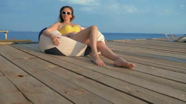 Attractive young woman in a yellow swimsuit relaxes sitting in an armchair bag on a wooden veranda above the sea. — Stock Footage Attractive young woman in a yellow swimsuit relaxes sitting in an armchair bag on a wooden veranda above the sea. Royalty Free Stock Footage