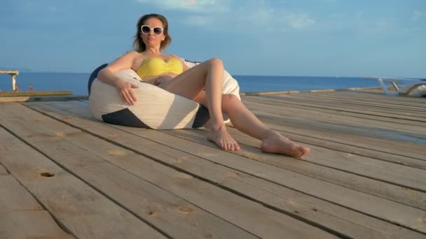 Attractive young woman in a yellow swimsuit relaxes sitting in an armchair bag on a wooden veranda above the sea. — Stock Video