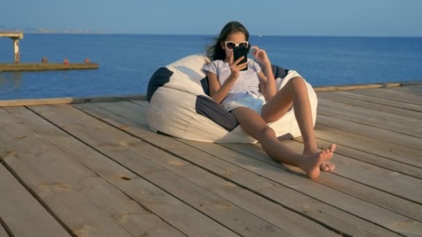 Menina adolescente na moda em óculos de sol descansando, sentado em uma cadeira saco em um terraço de madeira sobre o mar. usa seu smartphone — Vídeo de Stock