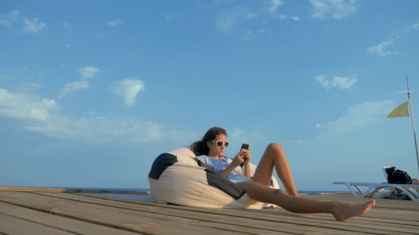 Adolescente de moda en gafas de sol descansando, sentado en una silla bolsa en una terraza de madera sobre el mar. utiliza su teléfono inteligente — Vídeo de stock