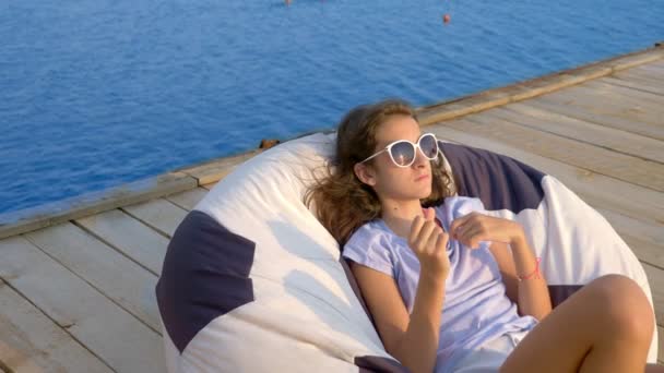 Adolescente de moda en gafas de sol descansando, sentado en una silla bolsa en una terraza de madera sobre el mar. utiliza su teléfono inteligente — Vídeo de stock