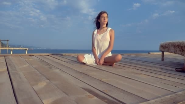 Menina adolescente em um vestido branco com cabelos longos olha pensativamente para o mar, estando em um terraço de madeira acima do mar. vento desenvolve cabelo — Vídeo de Stock