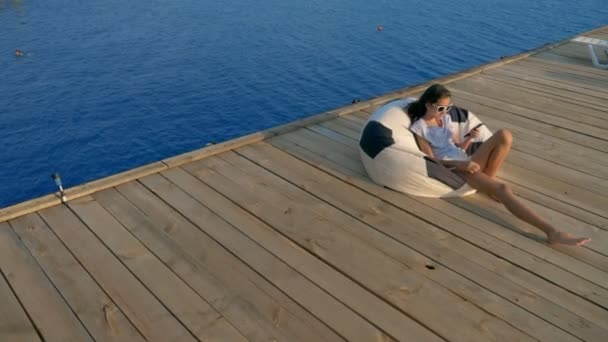 Adolescente de moda en gafas de sol descansando, sentado en una silla bolsa en una terraza de madera sobre el mar. utiliza su teléfono inteligente — Vídeos de Stock