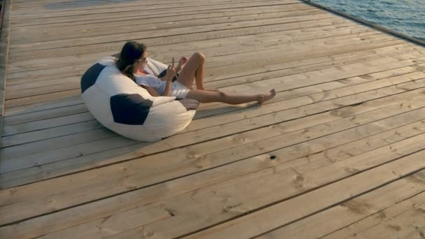 Модна дівчина-підліток в сонцезахисних окулярах відпочиває, сидячи в мішкуватому кріслі на дерев'яній терасі над морем. використовує свій смартфон — стокове відео