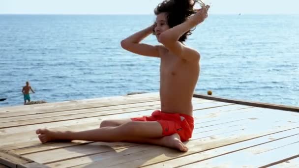 Kıvırcık saçlı genç çocuk deniz üzerinde ahşap bir terasta oturan deniz manzara fonuna karşı denize bakar — Stok video