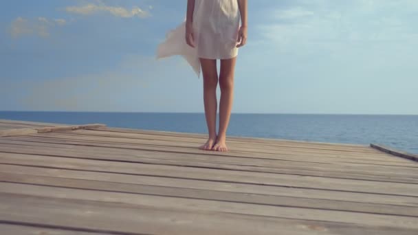 Menina adolescente em um vestido branco com cabelos longos olha pensativamente para o mar, estando em um terraço de madeira acima do mar. vento desenvolve cabelo — Vídeo de Stock