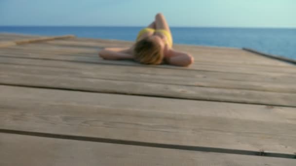 Mooie slanke vrouw met prachtige borsten in een helder roze badpak leugens en zonnebaden op een houten terras boven de zee. — Stockvideo