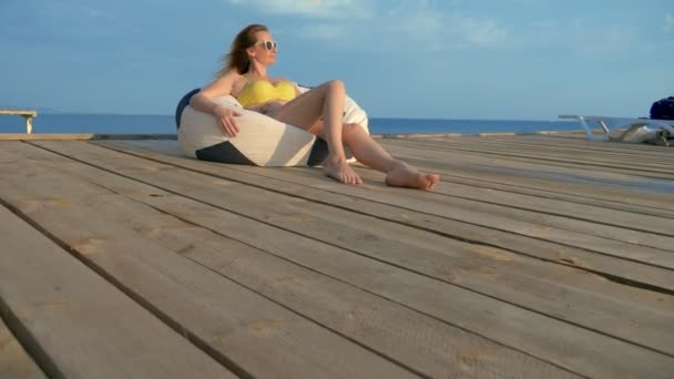 Atractiva mujer joven en un traje de baño amarillo se relaja sentado en una bolsa de sillón en una terraza de madera sobre el mar . — Vídeos de Stock