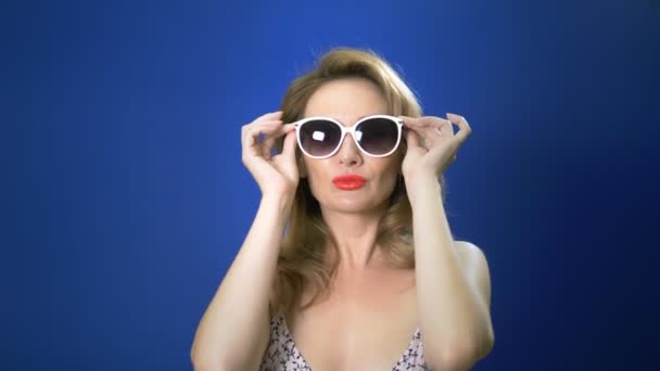 Pin-up-Girl-Konzept. Charmante Frau, die in die Kamera schaut und sich eine Sonnenbrille aufsetzt. blauer Hintergrund. Kopierraum — Stockvideo
