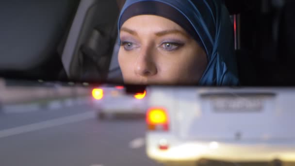 Reflejo de una mujer en un hiyab en un espejo retrovisor de un coche mientras conduce en un atasco de tráfico por la noche. Mujer musulmana conduciendo un coche . — Vídeos de Stock