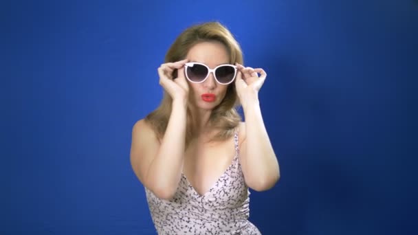 Pin-up-Girl-Konzept. Charmante Frau, die in die Kamera schaut und sich eine Sonnenbrille aufsetzt. blauer Hintergrund. Kopierraum — Stockvideo