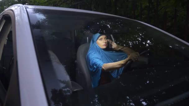 Zicht door de voorruit van een auto. Moslim vrouw in blauwe hijab praten op de telefoon tijdens het rijden van een auto. — Stockvideo