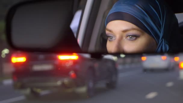 Reflet d'une femme dans un hijab dans un rétroviseur d'une voiture en conduisant dans un embouteillage le soir. Femme musulmane conduisant une voiture . — Video