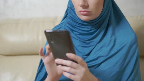 Bella donna musulmana in hijab blu utilizzando un telefono cellulare seduto su un divano — Video Stock