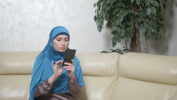 Piękna muzułmańska kobieta w niebieskim hidżab za pomocą telefonu komórkowego siedzi na kanapie — Wideo stockowe