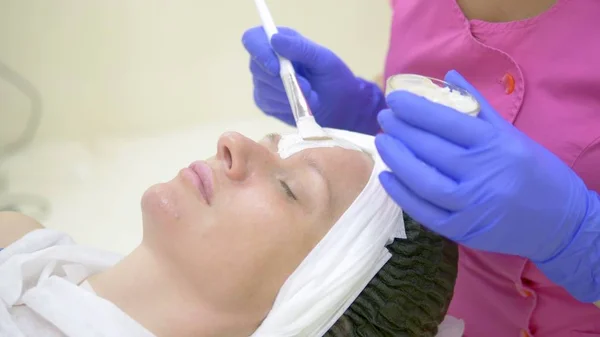 The concept of cosmetology. Close-up of a beautician applying a white mask with a brush on a womans face.