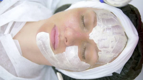 The concept of cosmetology. Close-up of a beautician applying a white mask with a brush on a womans face.