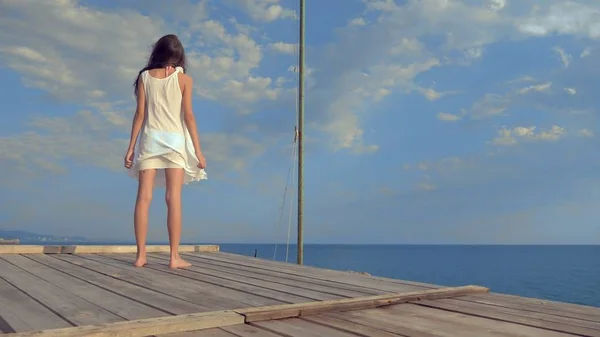 Adolescente en un vestido blanco con el pelo largo se ve pensativamente en el mar, estar en una terraza de madera sobre el mar. viento desarrolla el cabello — Foto de Stock