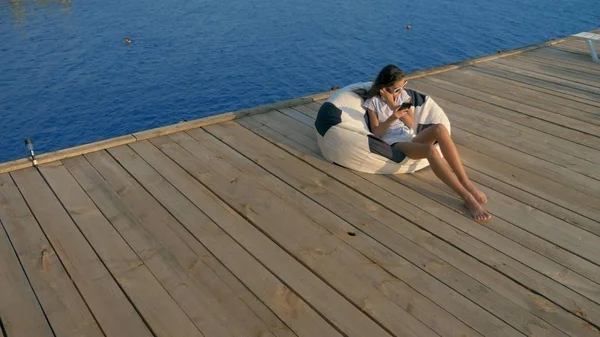 Gadis remaja modis berkacamata hitam beristirahat, duduk di kursi tas di teras kayu di atas laut. menggunakan smartphone-nya — Stok Foto