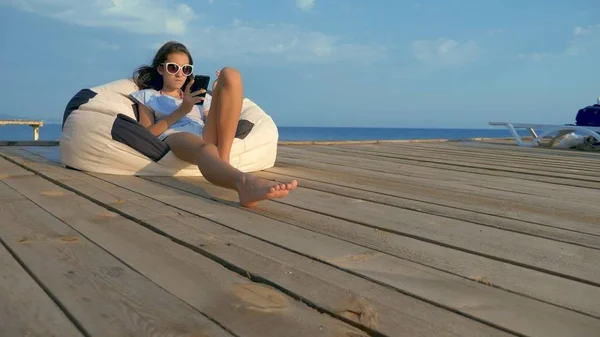 Modisches Teenager-Mädchen mit Sonnenbrille, das in einem Sackstuhl auf einer Holzterrasse über dem Meer sitzt. nutzt sein Smartphone — Stockfoto