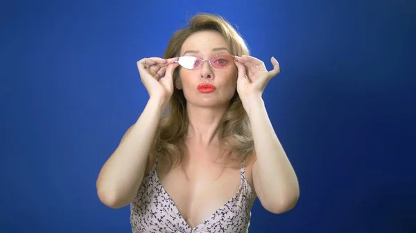 Pin-up-Girl-Konzept. Charmante Frau, die in die Kamera schaut und eine rosa Brille aufsetzt. blauer Hintergrund. Kopierraum — Stockfoto