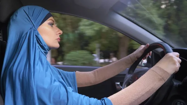 Hermosa mujer musulmana en hiyab azul conduciendo un coche. paseos durante el día por las calles de la ciudad . — Foto de Stock