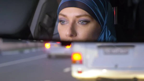 Reflejo de una mujer en un hiyab en un espejo retrovisor de un coche mientras conduce en un atasco de tráfico por la noche. Mujer musulmana conduciendo un coche . — Foto de Stock