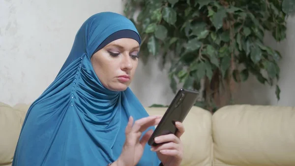 Hermosa mujer musulmana en hijab azul usando un teléfono móvil sentado en un sofá — Foto de Stock